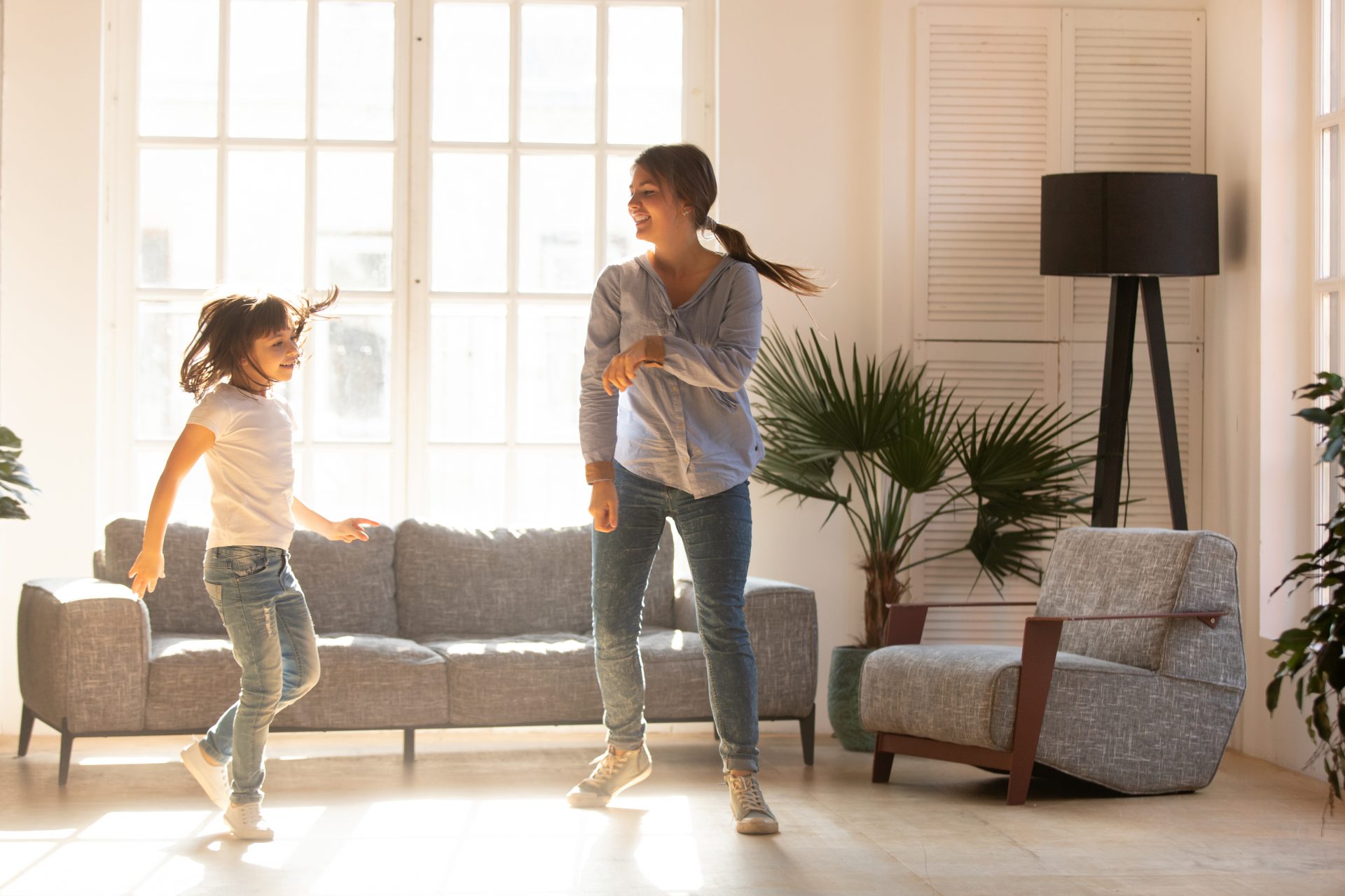 Mother and daughter dancing in a bright living room with large energy-efficient windows, enjoying natural light and a cozy home atmosphere.